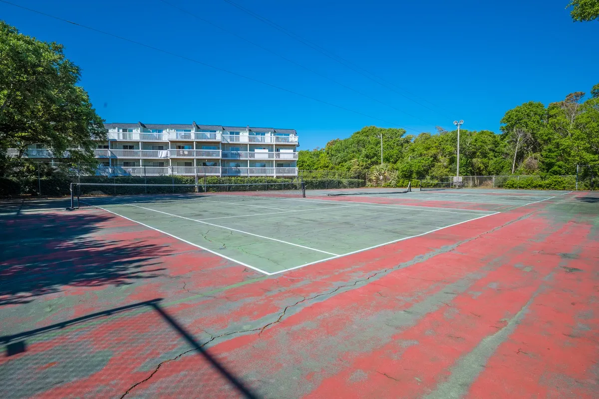 A well-maintained outdoor tennis court surrounded by lush greenery, with the resort building visible in the background. The court invites both competitive matches and casual play in a scenic outdoor setting.