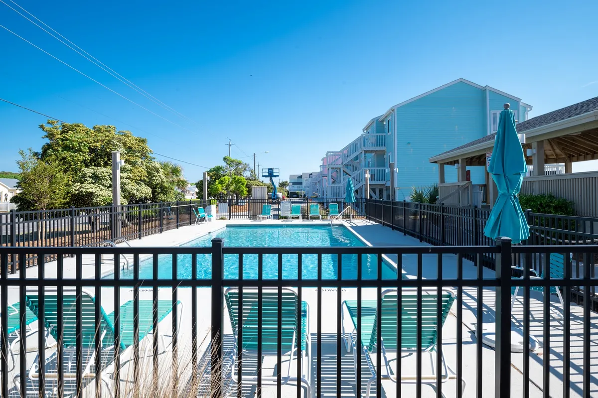 A serene exterior shot featuring a sparkling swimming pool bordered by comfortable lounge chairs and umbrellas, all enclosed by tasteful fencing. The ambiance is relaxing, with space for both sunbathing and evening gatherings, and hints of sunset light create a welcoming atmosphere.