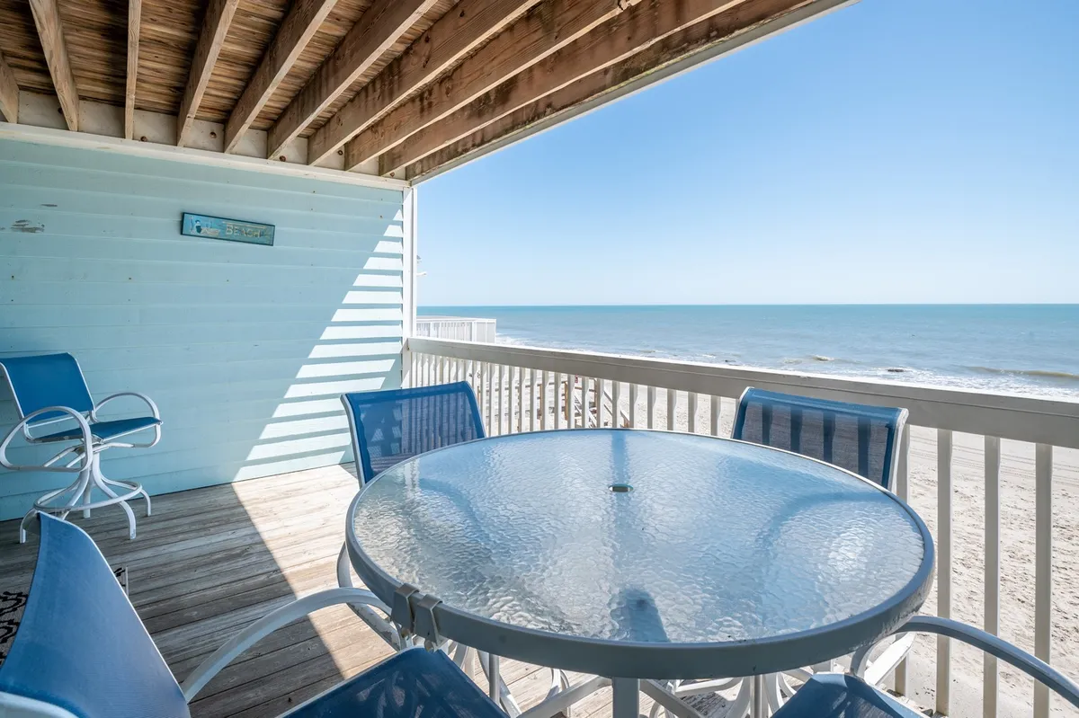 This exterior image showcases a glass-top table surrounded by blue chairs on a deck, overlooking a sweeping ocean view. The serene setting is ideal for outdoor dining, morning coffee, or sunset relaxation, with the horizon in clear sight.