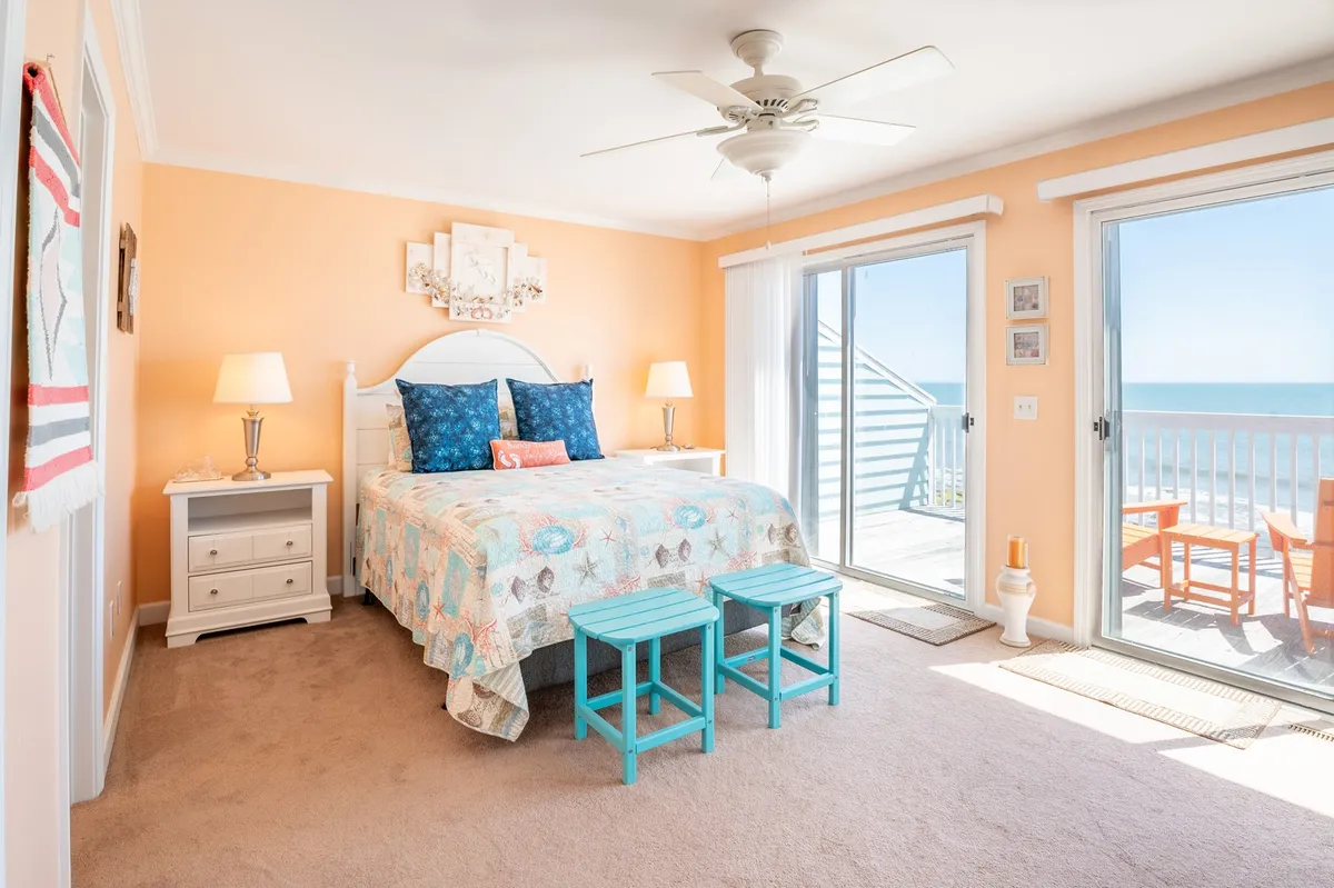 The image shows a serene bedroom featuring a plush bed adorned with colorful, inviting décor. Large windows frame stunning ocean views, filling the space with natural light and a tranquil atmosphere. A cozy reading nook is situated by the window, and the balcony is visible, hinting at the experience of enjoying morning coffee or evening drinks while overlooking the waves.