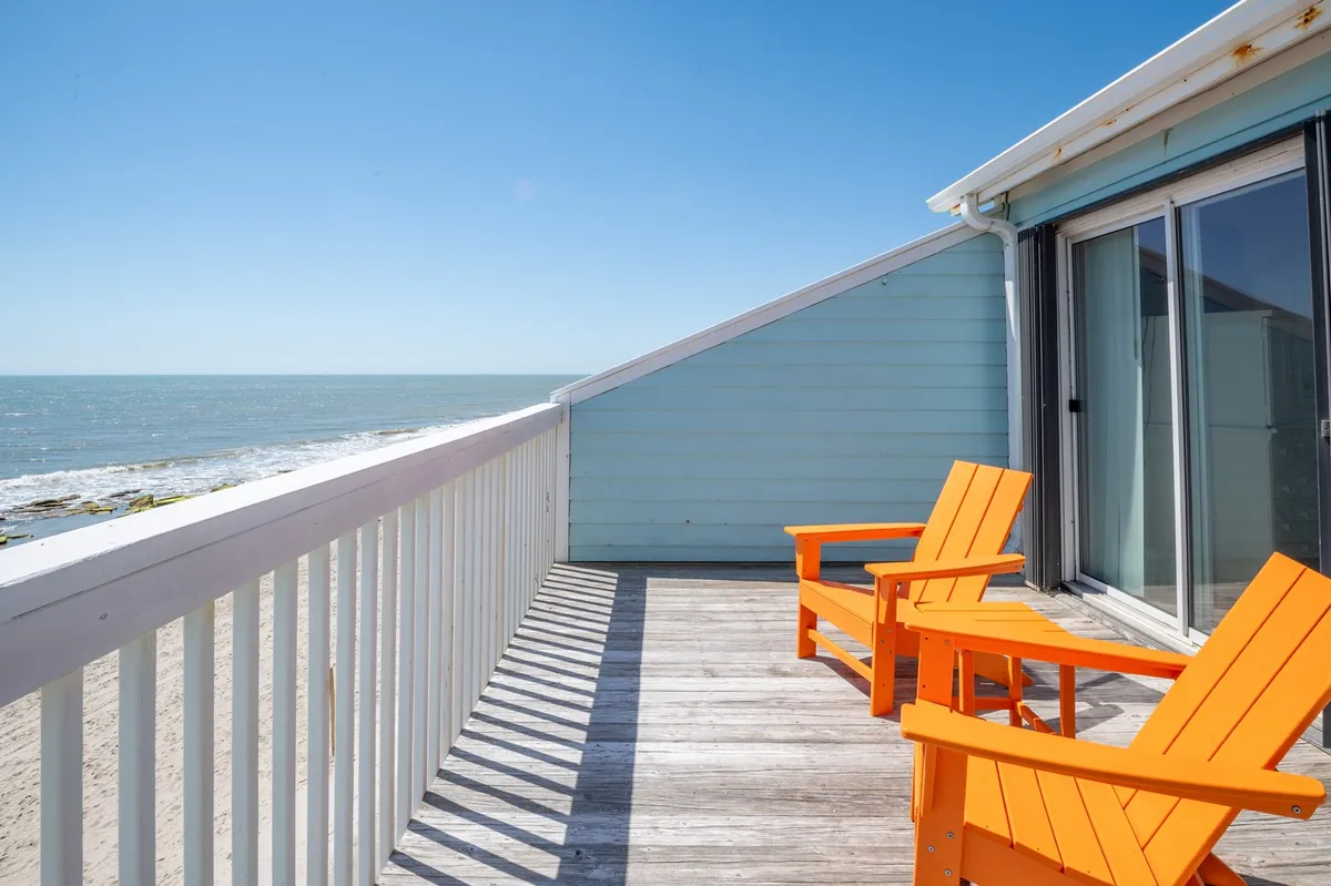 This image shows two orange Adirondack chairs on a wooden deck overlooking the ocean. The scene radiates peace and warmth, ideal for sipping morning coffee, enjoying sunset cocktails, or simply soaking up the coastal atmosphere. The connection between interior comfort and outdoor beauty is strongly conveyed.