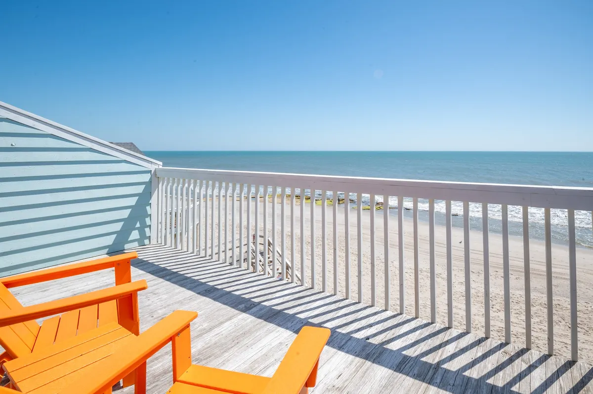Orange Adirondack chairs on a wooden deck face out toward an uninterrupted ocean view and clear blue sky, creating a tranquil setting for lounging, reflection, or soaking in the ocean breeze.