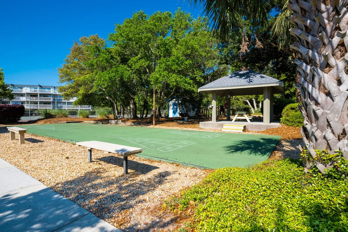 A peaceful outdoor communal area featuring a green playing surface, a gazebo, benches, and surrounding trees. The space invites group games, gatherings, and moments of relaxation in a natural setting.