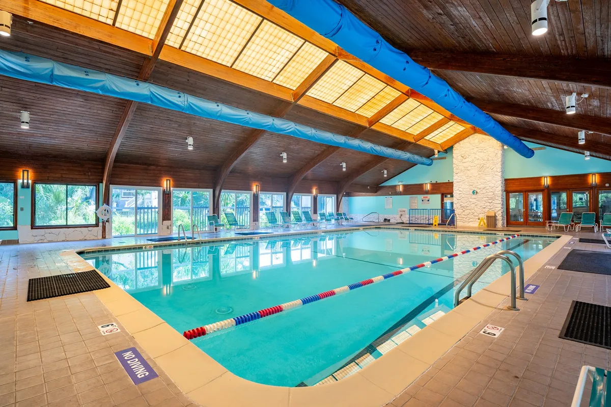 An indoor swimming pool area featuring wooden beams, expansive windows letting in natural light, and poolside lounge chairs. The space is bright and inviting, perfect for year-round swimming and social gatherings regardless of weather.