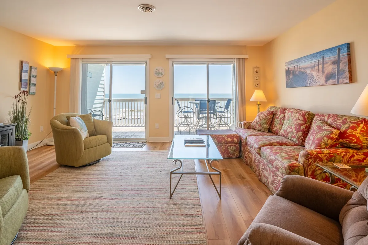 A bright, cheerful living room with large windows framing an ocean view. The space features a colorful floral couch, wooden coffee table, and comfortable seating, creating an inviting spot to relax, entertain, or enjoy the scenery. The combination of natural light and vibrant decor adds to the sense of comfort and togetherness.