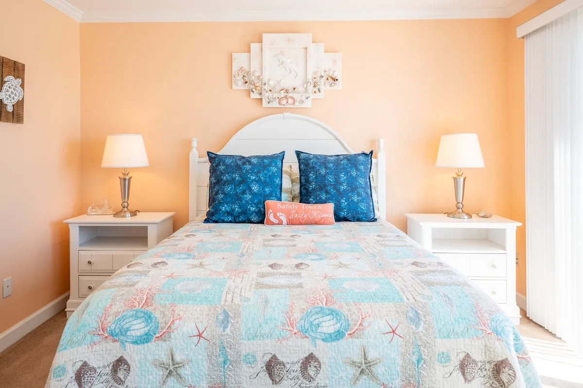 A serene bedroom with a queen-sized bed adorned with decorative pillows, flanked by nightstands with soft-glowing lamps, and calming wall art above the headboard. Gentle natural light filters through an unseen window, bathing the room in a warm, restful glow.