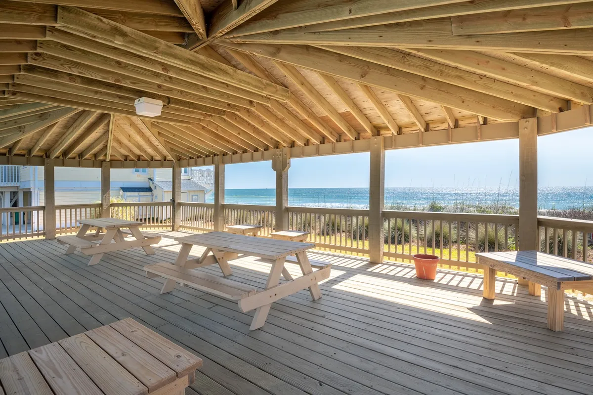 This image captures wooden picnic tables under shelter with a sweeping ocean view in the background. The scene is set for family gatherings, sunset dinners, or relaxing with a morning coffee, all with the ocean as a backdrop.