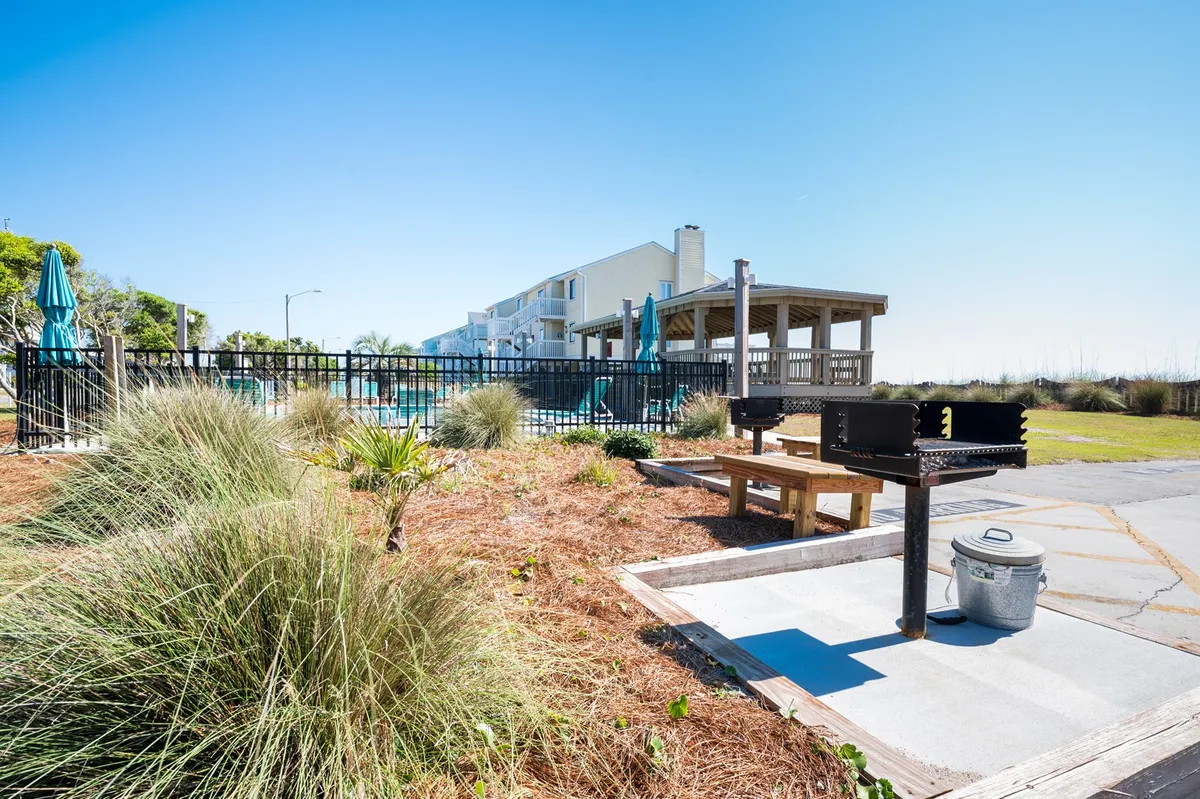 A vibrant outdoor scene showcasing a barbecue grill and picnic table beside a glistening pool surrounded by green umbrellas and manicured grass. The area is ideal for casual socializing, outdoor dining, and relaxed afternoons.